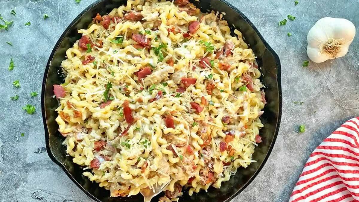 A cast iron skillet filled with creamy pasta topped with chopped bacon, herbs, and grated cheese. A spoon rests inside. A striped red and white towel and a head of garlic lie beside the skillet on a textured surface.