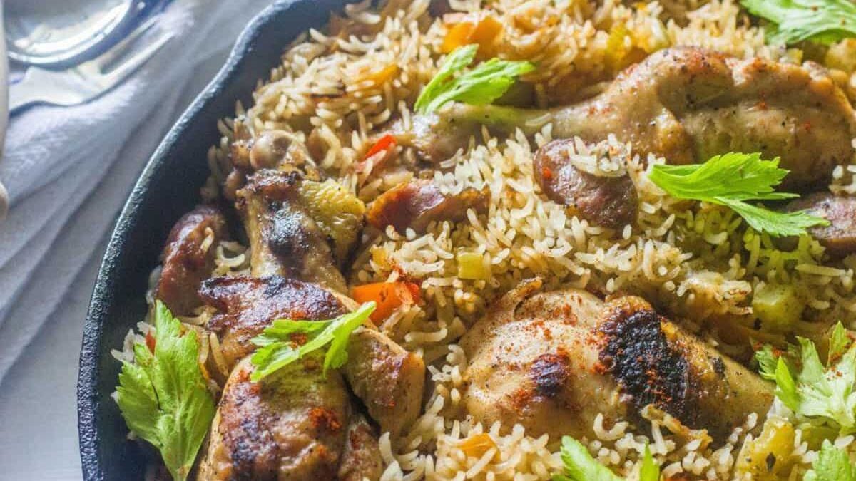 A close-up of a skillet filled with a cooked rice dish, featuring fluffy basmati rice mixed with vegetables and topped with seasoned chicken drumsticks. Garnished with fresh green herbs, the dish looks flavorful and appetizing. A spoon and napkin are visible in the background.