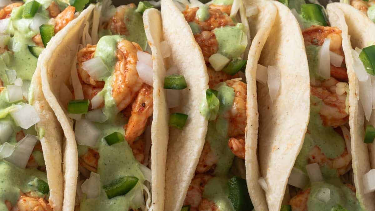 Close-up of tacos filled with shrimp and topped with diced onions, chopped jalapeños, and a drizzle of green sauce. The tacos are in soft corn tortillas arranged in a row.