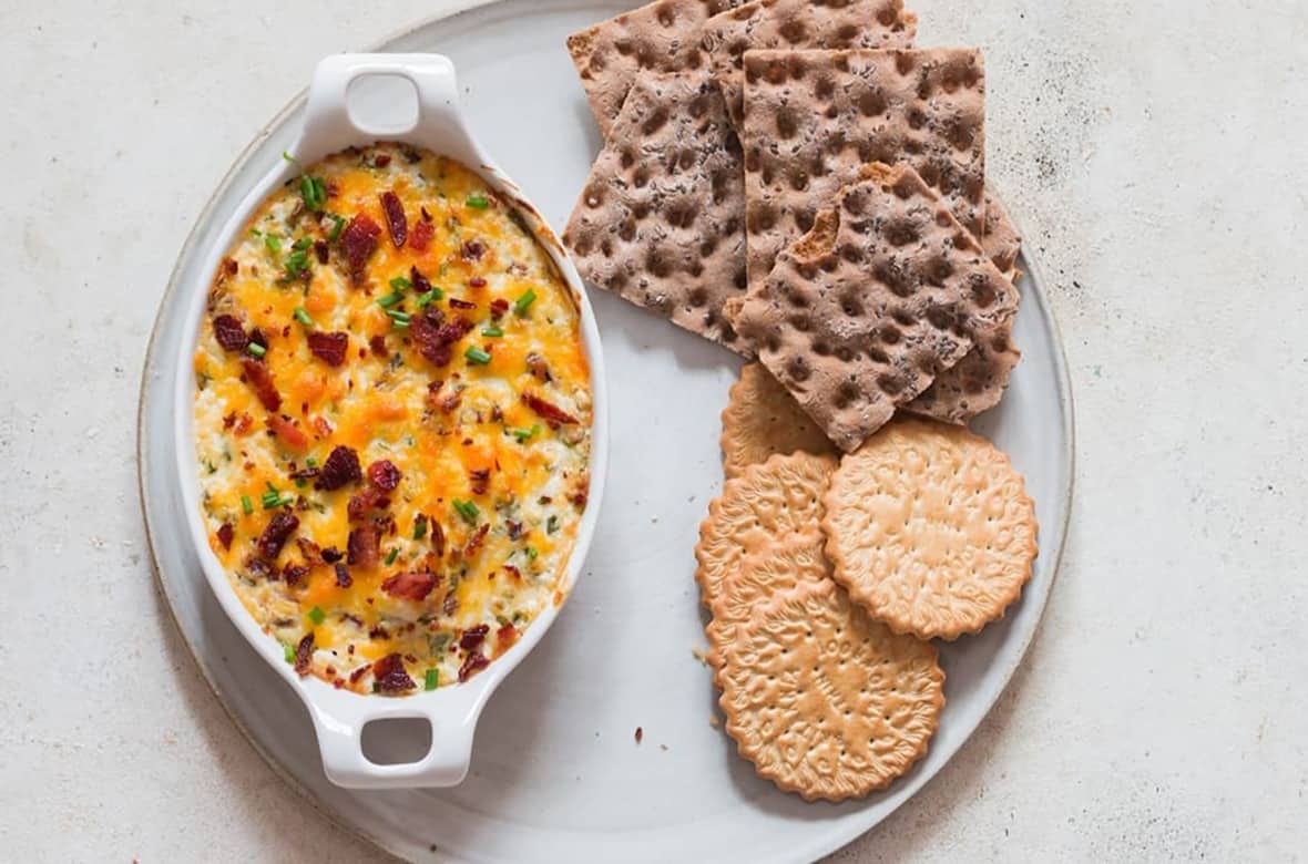 A baked cheesy dip topped with herbs and bacon in a white oval dish on a round plate, accompanied by crispy whole-grain crackers and round biscuits.