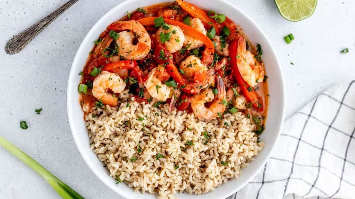 A white bowl filled with brown rice and a shrimp and vegetable stir-fry, topped with sliced red bell peppers and green onions, sits on a light-colored surface with a spoon and a lime wedge in the background. A white checkered napkin is partially visible to the side.