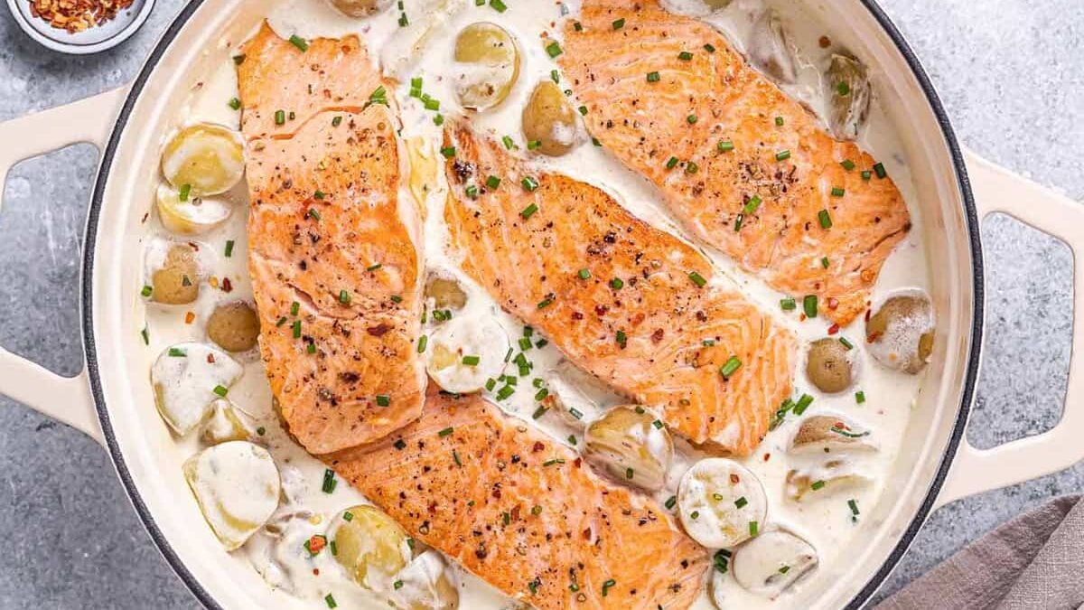 A creamy dish featuring four seasoned salmon fillets garnished with chives in a rich, white sauce with halved baby potatoes. A small bowl of red chili flakes is visible in the upper left corner. The dish is presented in a white skillet on a gray surface.
