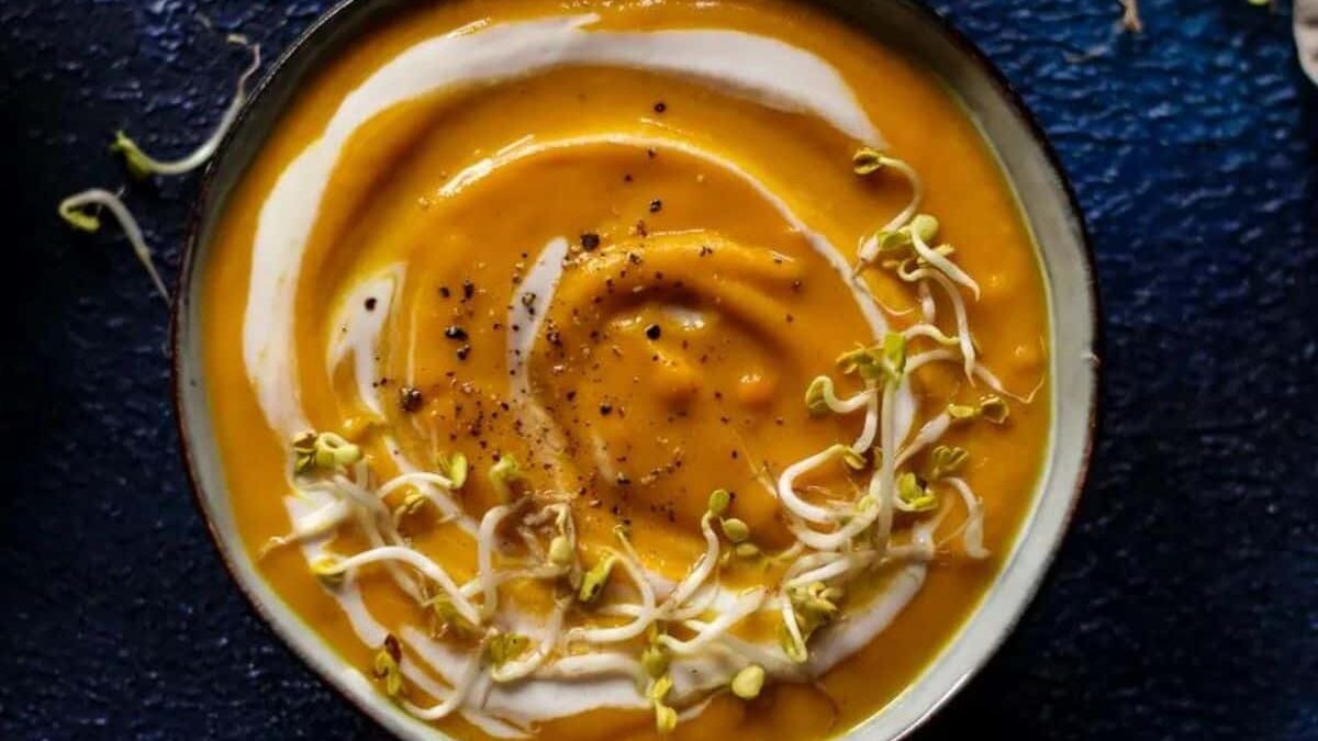 A bowl of creamy orange soup garnished with a swirl of white cream, black pepper, and small green sprouts, sits on a dark blue, textured surface. The soup appears smooth and appetizing.
