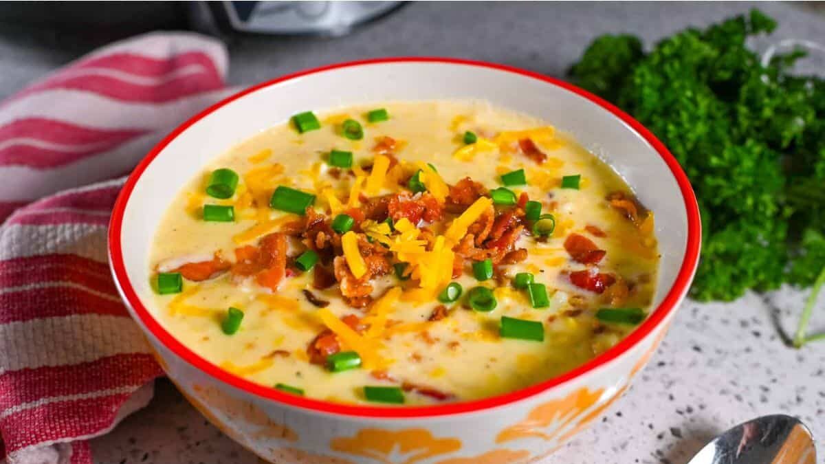A bowl of creamy potato soup topped with crispy bacon, shredded cheddar cheese, and chopped green onions. The bowl rests on a speckled countertop next to a red-striped napkin and fresh parsley. A spoon is placed beside the bowl.