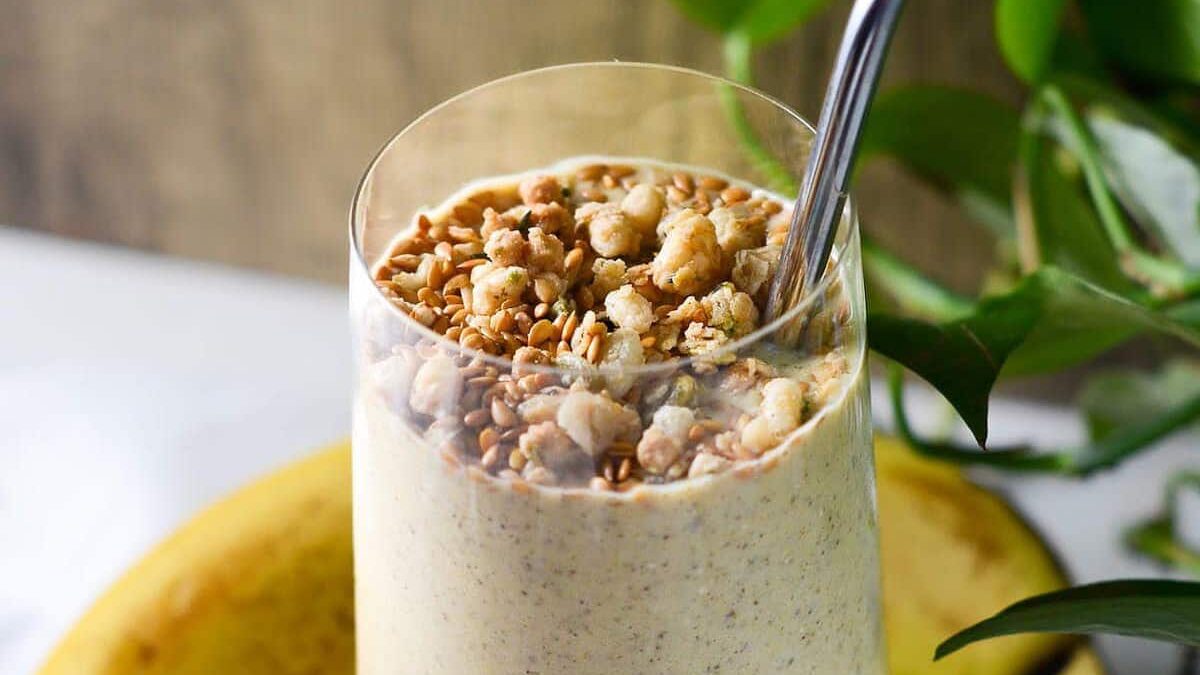 A close-up of a smoothie topped with granola pieces and a metal straw in a clear glass. In the background, there are partially visible green leaves and a ripe banana.