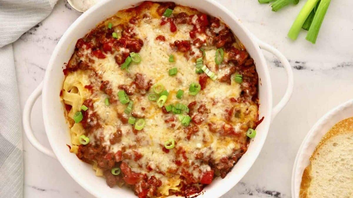 A round, white dish filled with baked pasta, topped with melted cheese, ground meat, tomato sauce, and garnished with chopped green onions, sits on a marble surface. A slice of bread and green onions are beside the dish. A white cloth is partially visible on the side.
