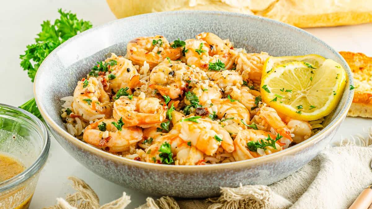 A bowl of shrimp scampi on a bed of rice, garnished with parsley and a lemon slice. A loaf of bread and a green garnish are in the background, alongside a plate with toasted bread. A small bowl with a sauce is next to the dish.