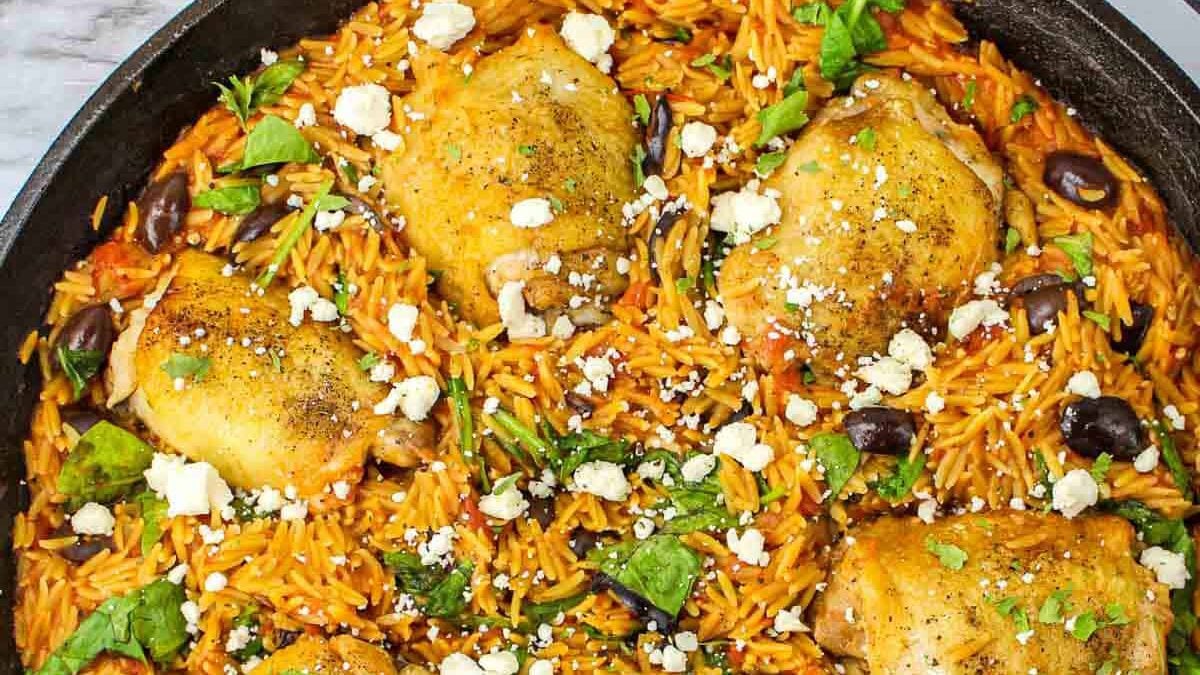 A close-up of a skillet filled with baked chicken thighs on a bed of seasoned orzo pasta. The dish is garnished with crumbled feta cheese, fresh herbs, and black olives, creating a vibrant and appetizing appearance.