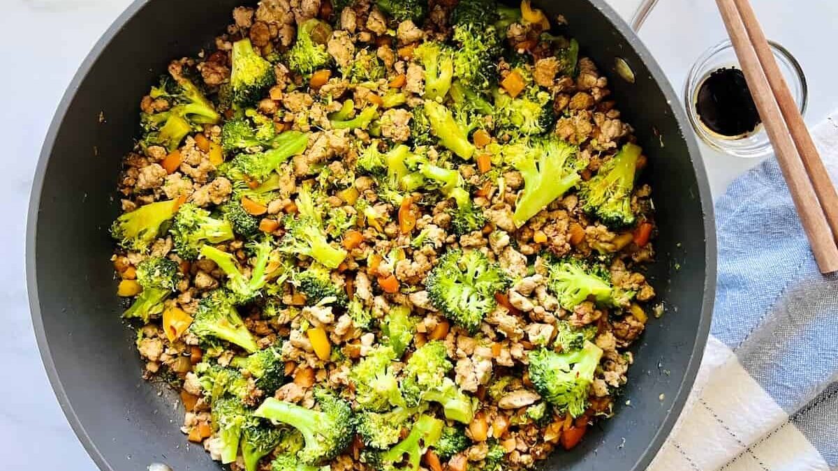A large pan filled with a colorful stir-fry dish consisting of broccoli florets, diced carrots, and ground meat. On the side, a small bowl of soy sauce and a pair of chopsticks sit on a striped cloth.