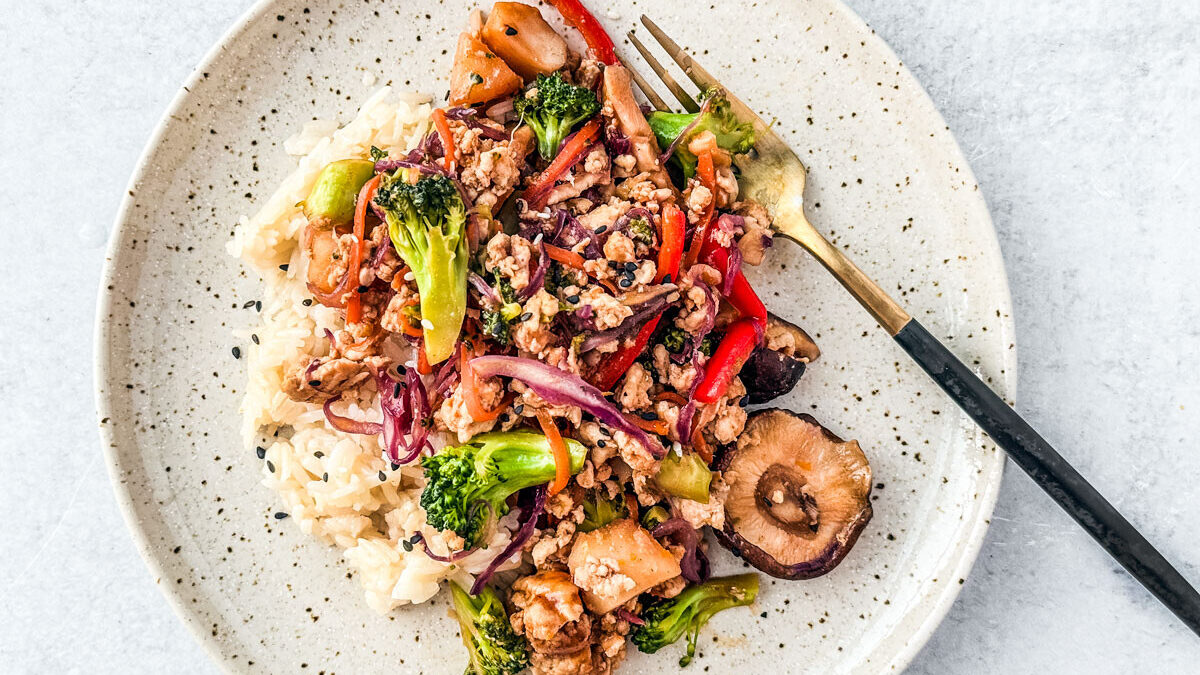 A plate of stir-fried vegetables with rice featuring broccoli, red bell peppers, mushrooms, and shredded cabbage. The dish is garnished with sesame seeds and served with a fork on a round, speckled plate.