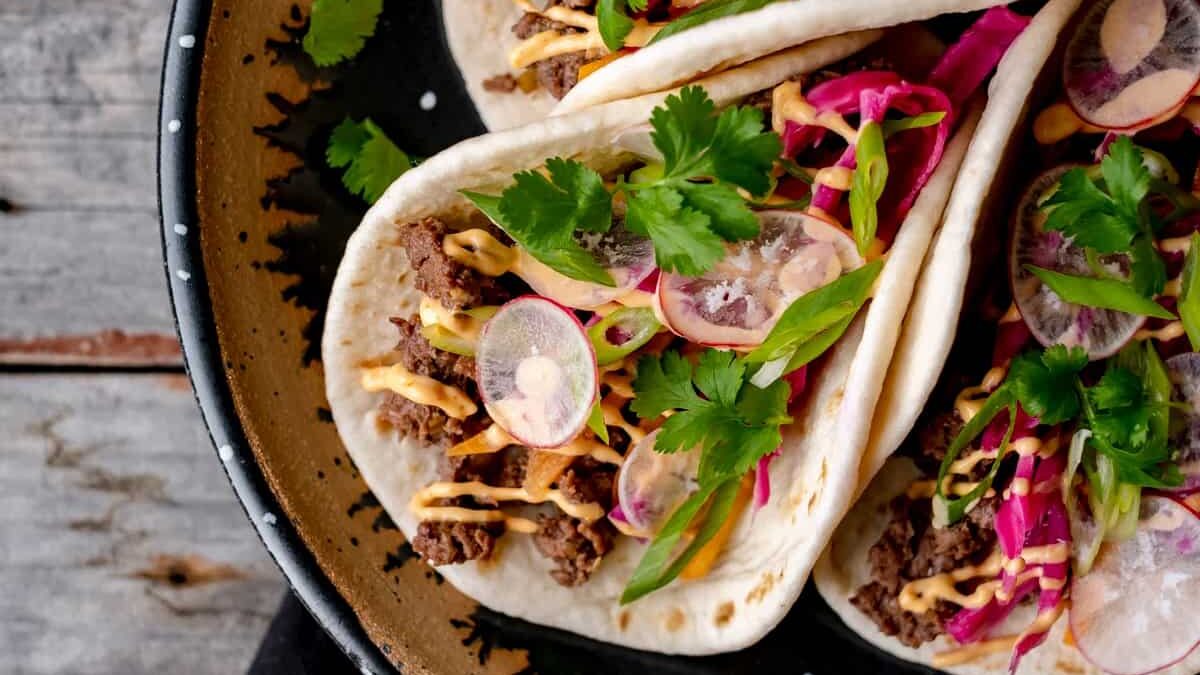 Three soft tacos on a black plate are filled with spiced meat, sliced radishes, cilantro, shredded purple cabbage, and drizzled with a creamy orange sauce. They are garnished with green onions and fresh herbs.