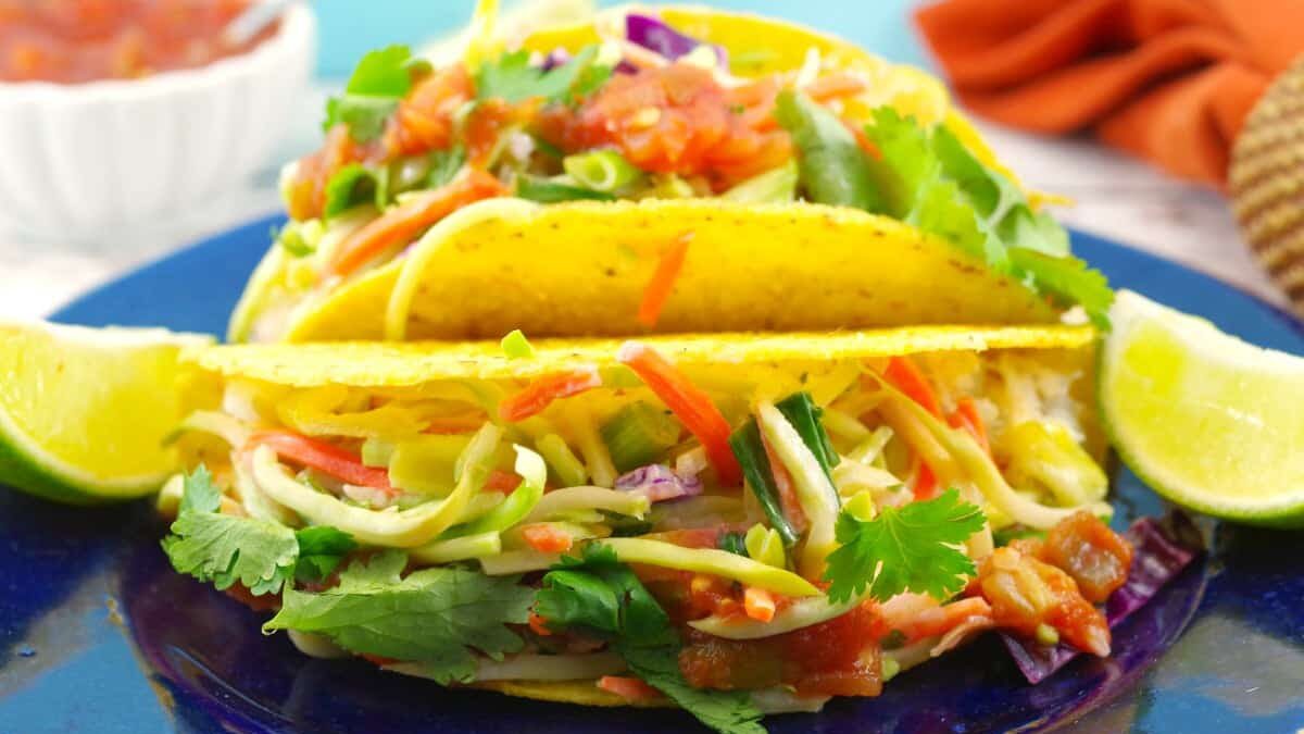 Two crispy tacos filled with vibrant veggies, garnished with cilantro, are served on a blue plate. Lime wedges are placed on the side, and a bowl of salsa in the background hints at fresh Mexican flavors.