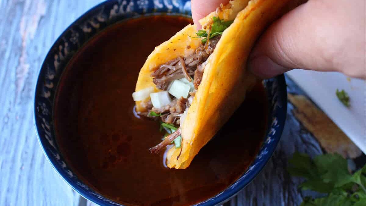 A hand holding a taco and dipping into a bowl with broth.
