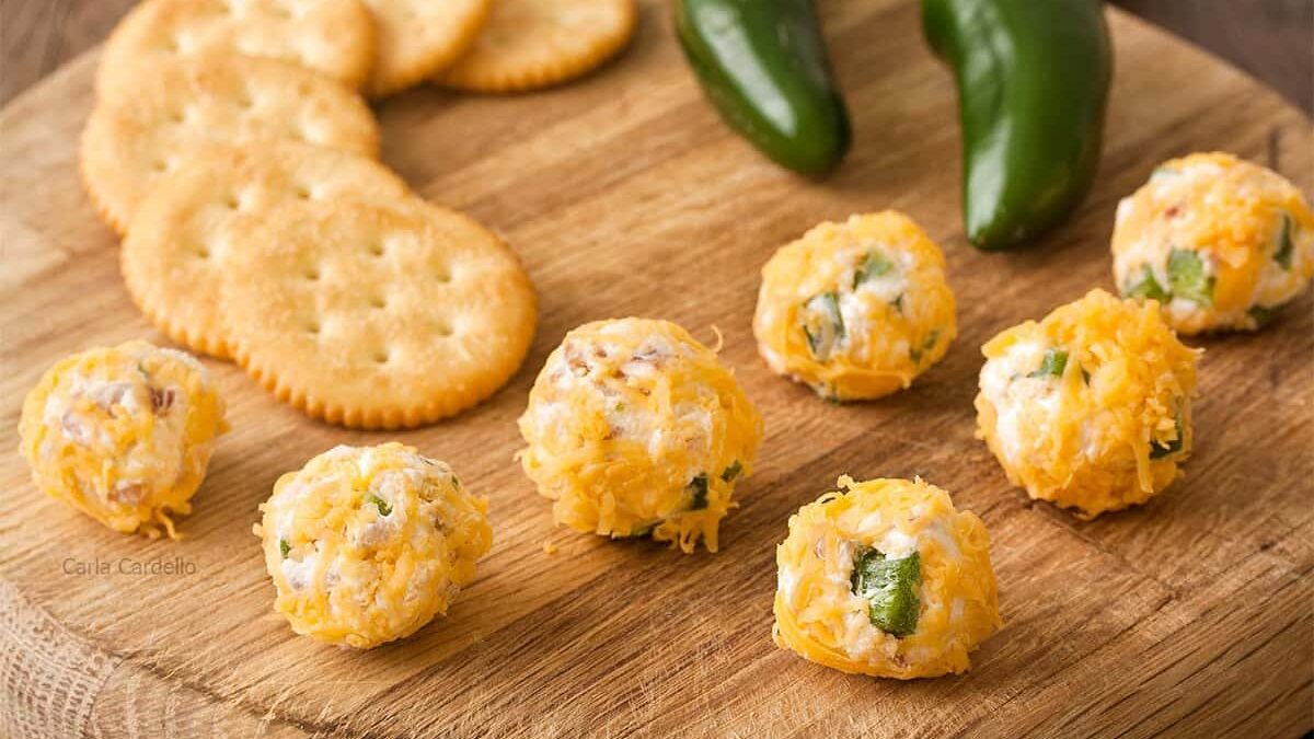 Cheese balls with jalapeño and herbs are arranged on a wooden board. They are accompanied by round crackers and two whole jalapeños in the background.