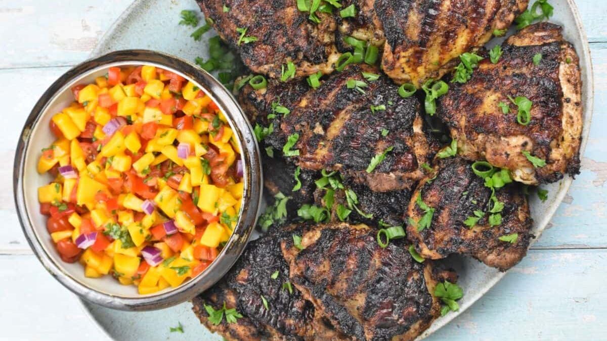 A plate of grilled chicken thighs garnished with chopped green herbs is served alongside a bowl of colorful mango salsa on a light blue wooden table. The salsa contains diced mango, red onion, tomatoes, and cilantro.