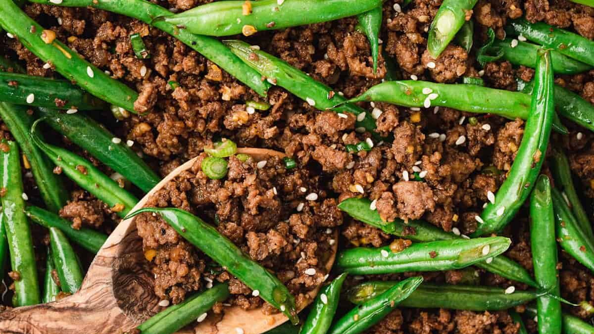 A wooden spoon scoops a mixture of cooked ground beef and fresh green beans, garnished with sesame seeds. The dish is seasoned and colorful, with vibrant green and rich brown tones.