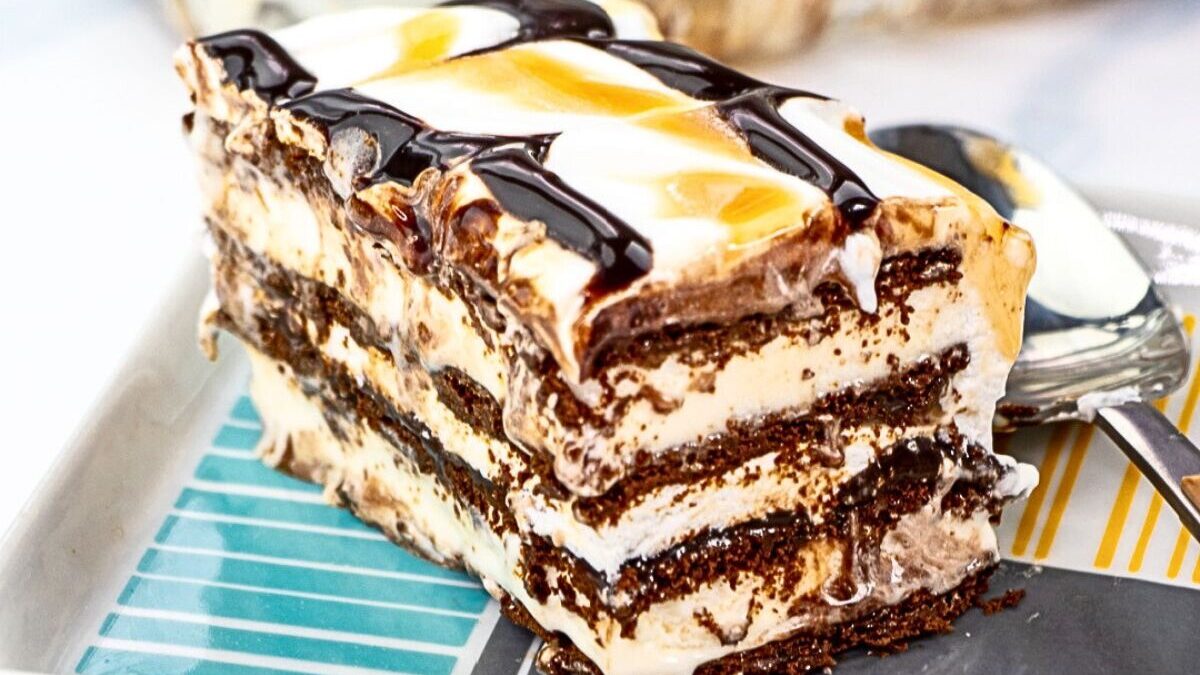 A slice of delectable ice cream cake boasts chocolate cookie layers, creamy white ice cream, and a drizzle of chocolate and caramel sauce on top. Perfectly presented on a rectangular plate with a spoon beside it, this is the epitome of delicious desserts.