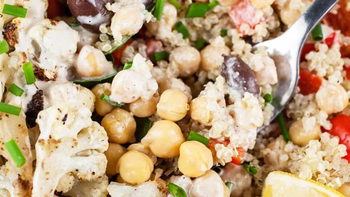 Close-up of a quinoa salad with chickpeas, roasted cauliflower, kalamata olives, red bell peppers, chopped chives, and a creamy dressing. A spoon is scooping the salad next to a lemon wedge.