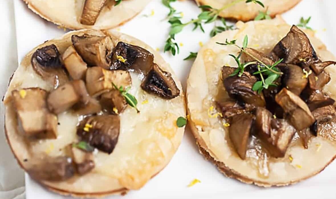 Four mini mushroom and cheese tarts garnished with fresh thyme are displayed on a white plate.