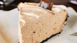 A slice of creamy pie with a chocolate crumb crust sits on a plate. The pie is topped with a small piece of chocolate. A fork is visible beside the plate.