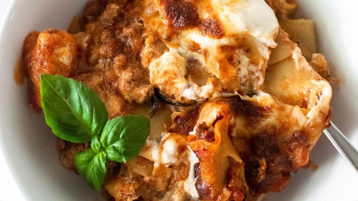 A close-up of a serving of lasagna in a white bowl, topped with melted cheese and a sprig of fresh basil, with a fork partially visible on the side.