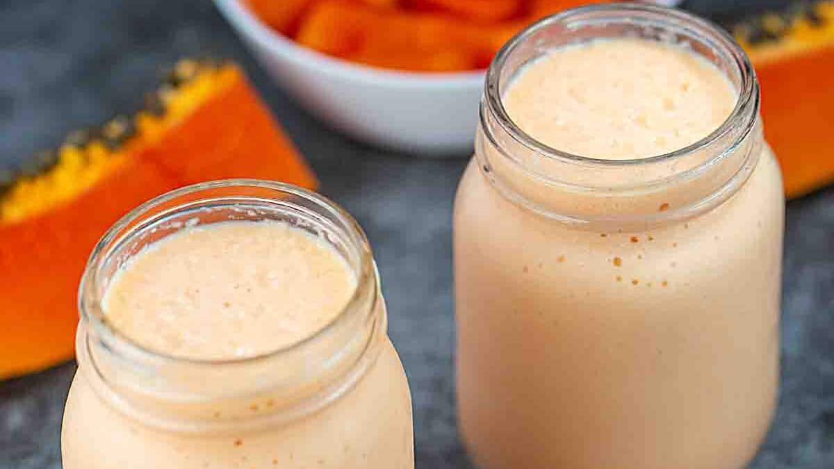 Two mason jars filled with creamy papaya milkshake sit on a gray surface. In the background, there's a white bowl containing orange papaya slices. The milkshake is light orange, matching the papaya's color.