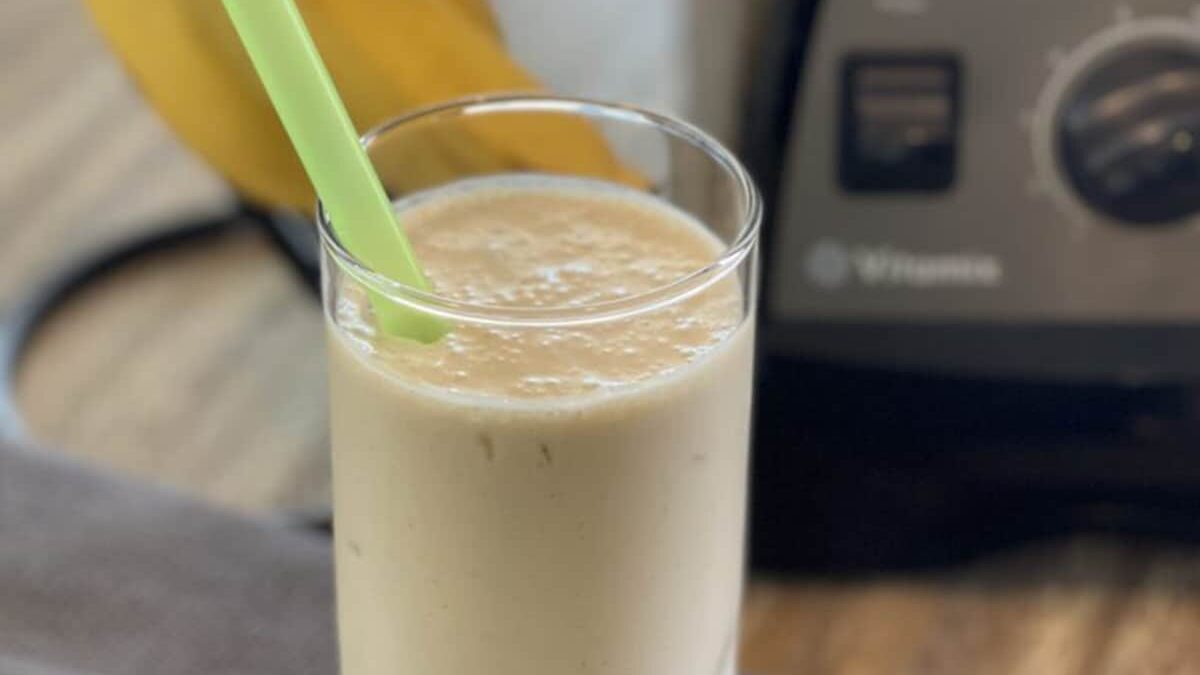 A creamy banana smoothie in a glass with a green straw, placed on a wooden table. A blender and a ripe banana are visible in the background.