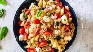 A black bowl filled with pasta salad containing cherry tomatoes, mozzarella balls, black olives, tuna, chopped herbs, and garnished with fresh basil leaves on a light-colored surface.