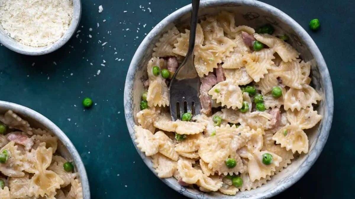 A bowl of farfalle pasta mixed with green peas, pieces of meat, and a creamy sauce. A fork rests inside the bowl. Grated cheese is sprinkled around, with a small bowl of extra cheese nearby.
