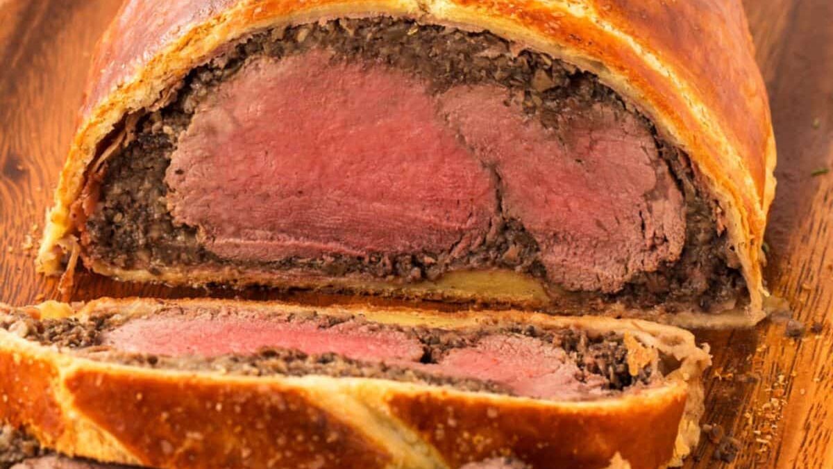 A close-up of a sliced beef Wellington on a wooden board. The pastry is golden brown and flaky, enclosing a layer of duxelles and medium-rare beef. Two slices are cut in front, revealing the cross-section.