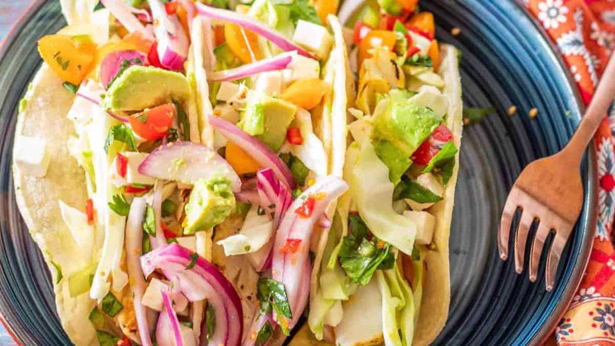 A plate of fresh, colorful tacos filled with avocado, cabbage, red onion, and diced tomatoes. The tacos are served on a dark blue plate with a fork resting beside them on a patterned red cloth.