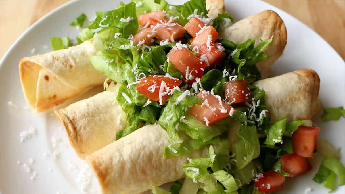 Potato flautas on a white plate topped with lettuce and tomatoes.