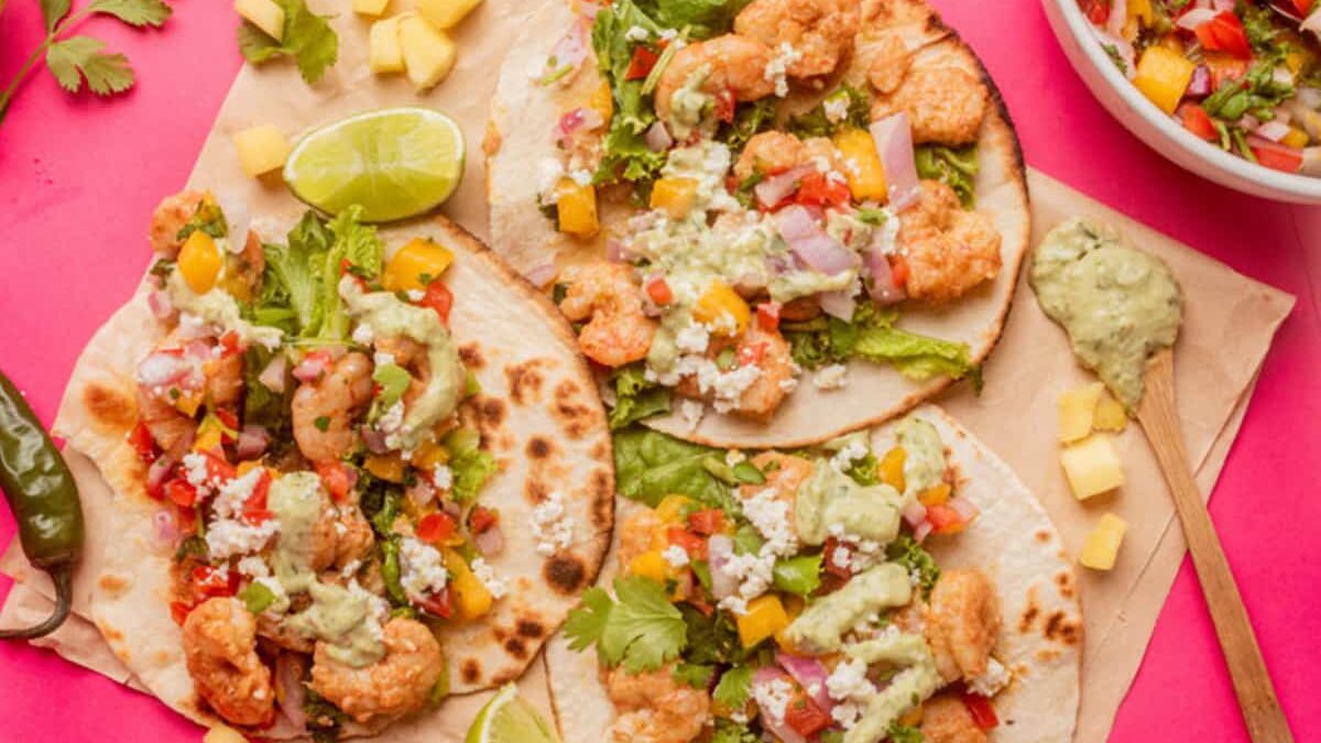 Three shrimp tacos on tortillas are topped with lettuce, diced tomatoes, red onions, cheese, and creamy sauce. Garnished with lime wedges and diced mango on a pink background, with a bowl of similar toppings nearby.