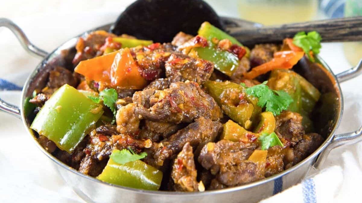 A bowl of stir-fried beef adorned with green peppers, tomatoes, and fresh cilantro, featuring a rich, savory sauce. A black serving spoon rests in the bowl, sitting on a white surface with a blue-striped cloth beside it.