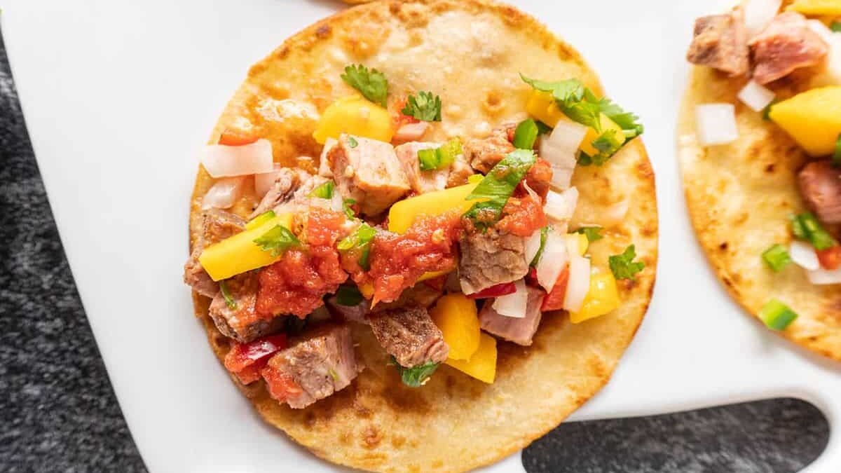 A taco on a white plate, filled with diced beef, mango chunks, red onions, cilantro, and topped with salsa. The taco shell is lightly toasted and golden brown.