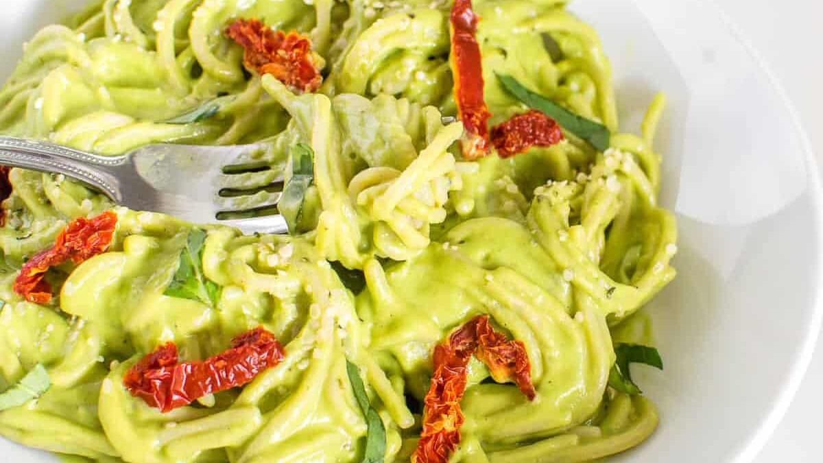 A bowl of spaghetti is covered in creamy green avocado sauce, garnished with sun-dried tomatoes and fresh basil. A fork is twirling the pasta, and the dish is presented on a white plate.