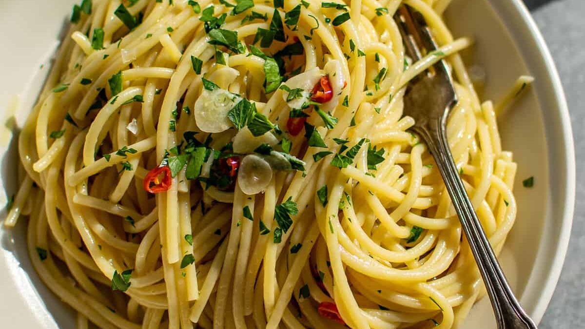 A plate of spaghetti aglio e olio garnished with chopped parsley, sliced garlic, and red pepper flakes. A fork is placed on the plate. The dish is light and glossy, reflecting natural light from the side.