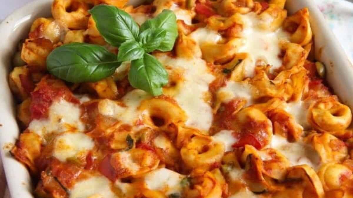 A close-up view of a baked pasta dish topped with melted cheese and garnished with fresh basil leaves. The pasta appears to be tortellini coated in a rich tomato sauce, served in a white baking dish.