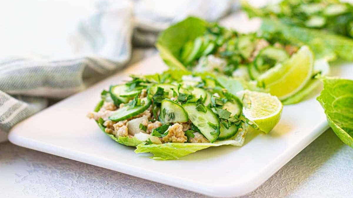 Two lettuce wraps filled with cooked ground meat, sliced cucumbers, and chopped herbs are displayed on a white rectangular plate. A lime wedge is placed beside the wraps. A striped cloth is in the background.