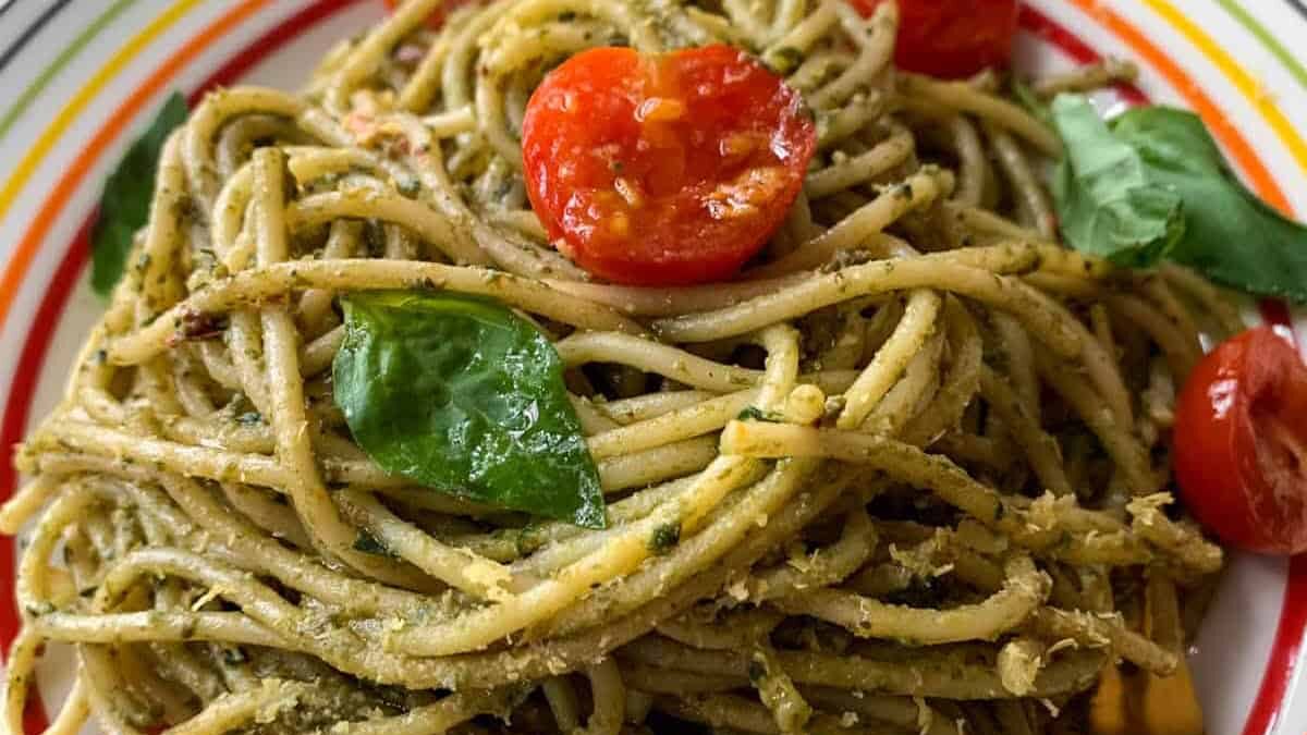 A plate of spaghetti coated in pesto sauce, topped with halved cherry tomatoes and fresh basil leaves. The dish is served on a colorful striped plate.
