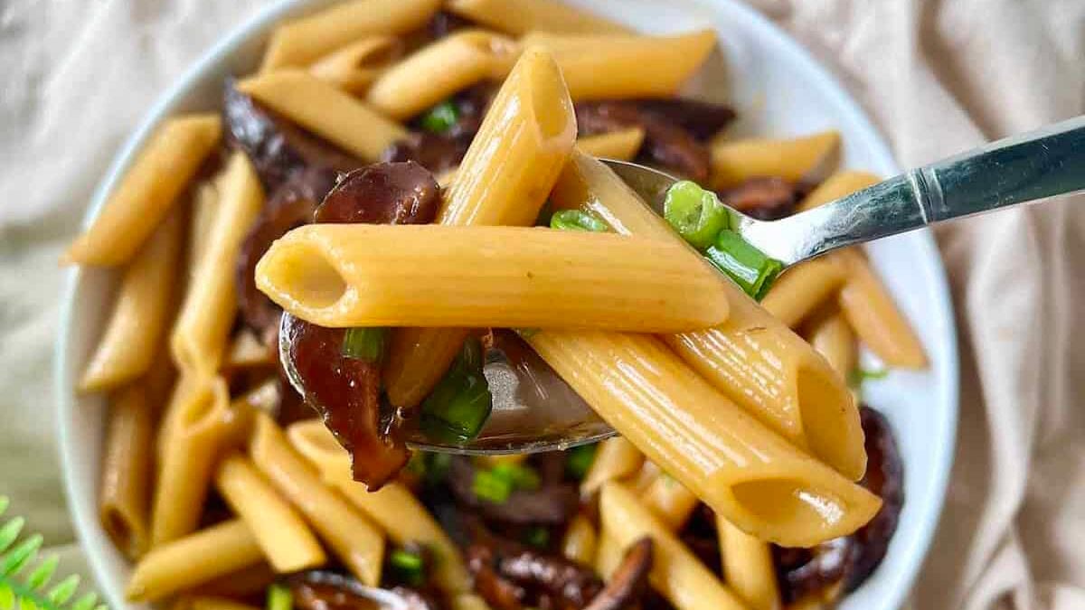 Close-up of a fork lifting penne pasta with mushroom slices and green onions from a bowl filled with more pasta and mushrooms. The background is a soft beige fabric.