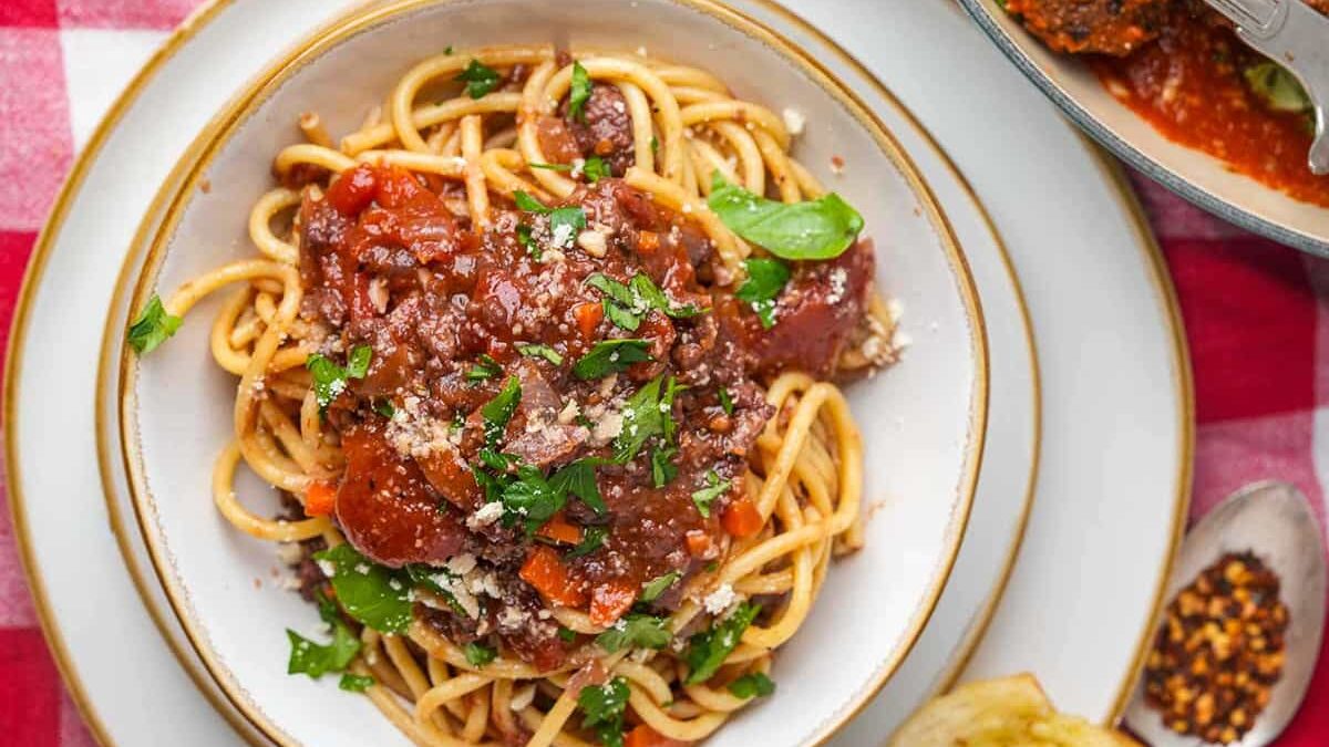 A plate of spaghetti topped with chunky bolognese sauce and fresh herbs, presented on a checkered red-and-white tablecloth. A spoon and a partially seen dish of extra sauce are nearby.