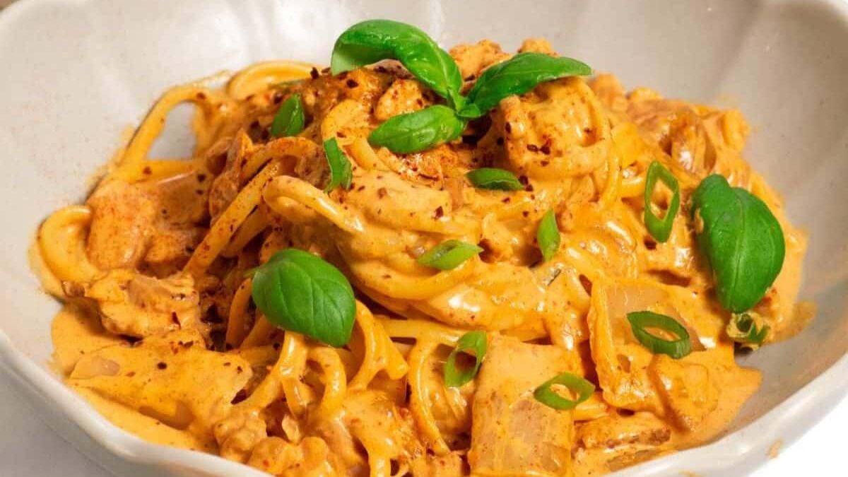 A bowl of creamy pasta topped with sliced chicken, green onions, and fresh basil leaves. The sauce is rich and orange, garnished with a sprinkle of red pepper flakes, all served in a white bowl.