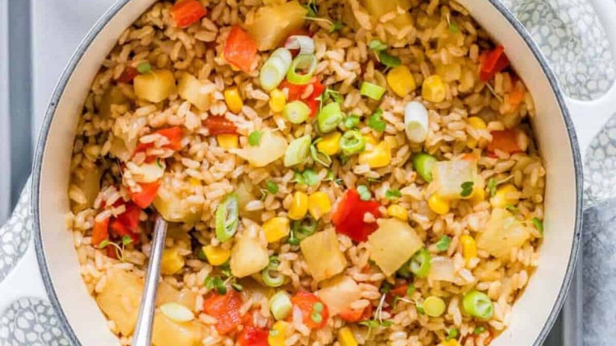 A pot filled with cooked rice mixed with diced red peppers, corn, pineapple chunks, and sliced green onions. A stainless steel spoon rests inside, ready for serving. The pot is placed on a decorative surface.