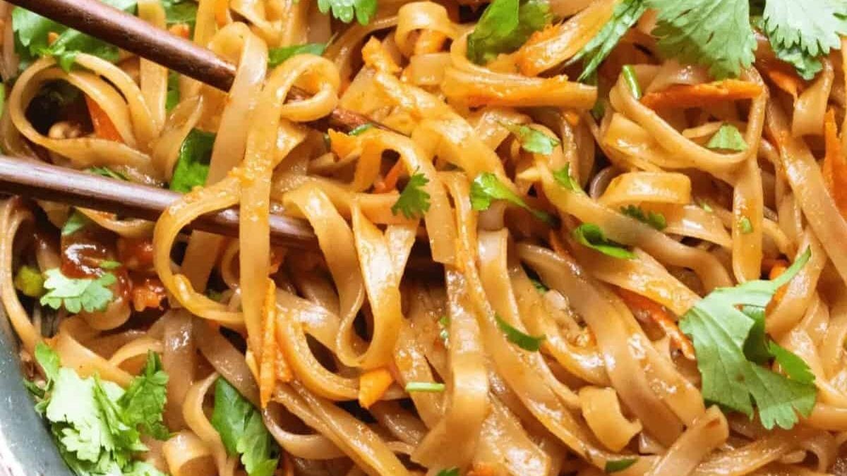 Close-up of a bowl of noodles mixed with shredded carrots, garnished with fresh cilantro. Two wooden chopsticks rest on the side. The dish has a glossy appearance, indicating it is well-seasoned or sauced.