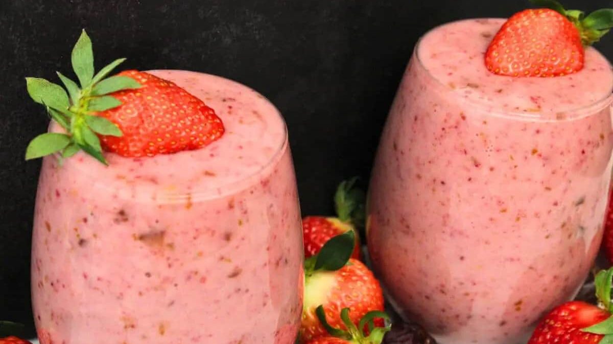 Two glasses filled with a creamy pink strawberry smoothie, each topped with a whole strawberry. Fresh strawberries surround the glasses, set against a dark background.