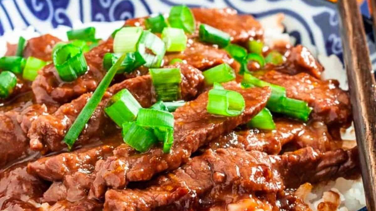 A close-up of a dish featuring slices of cooked beef topped with fresh green onions, served over white rice on a decorative blue and white plate. The beef appears glazed with a rich, savory sauce.