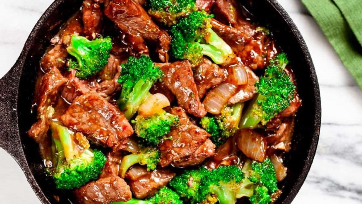 A skillet filled with beef pieces, broccoli florets, and onions cooked in a savory sauce. The dish is placed on a white marble surface with a green cloth partially visible on the side.