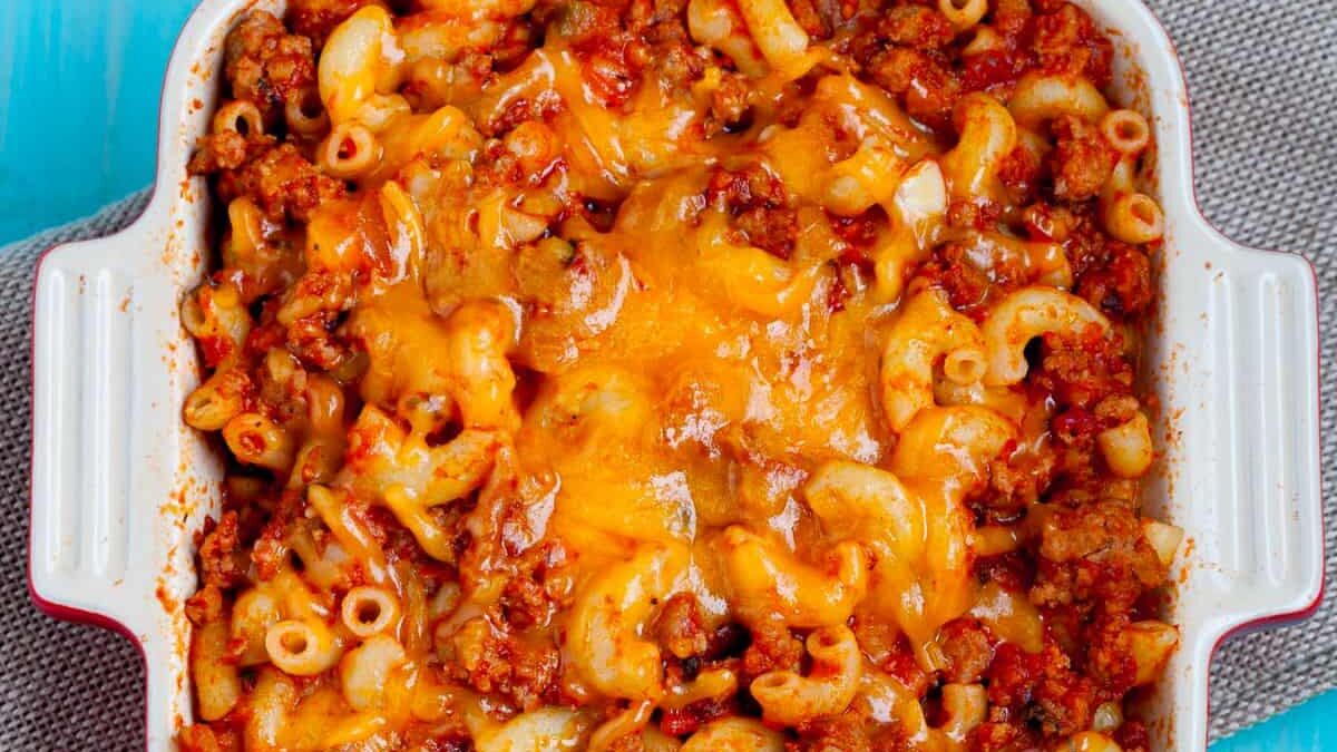 A baked macaroni casserole in a white square dish on a gray cloth. The dish contains elbow macaroni mixed with ground meat and topped with melted cheddar cheese, creating a bubbly and golden-brown crust.