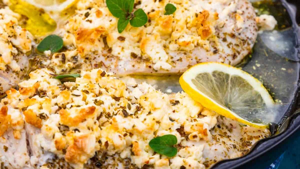 Close-up of seasoned baked chicken breasts topped with crumbled cheese and herbs in a pan. A slice of lemon and small green leaves garnish the dish, adding color and freshness.